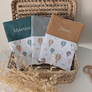 May include: Three personalized baby changing mats in a wicker basket. Each mat has a different color top: teal, gray, and tan. The mats are embroidered with the names "Marius", "Ed", and "Jean". The bottom portion features a hot air balloon pattern.