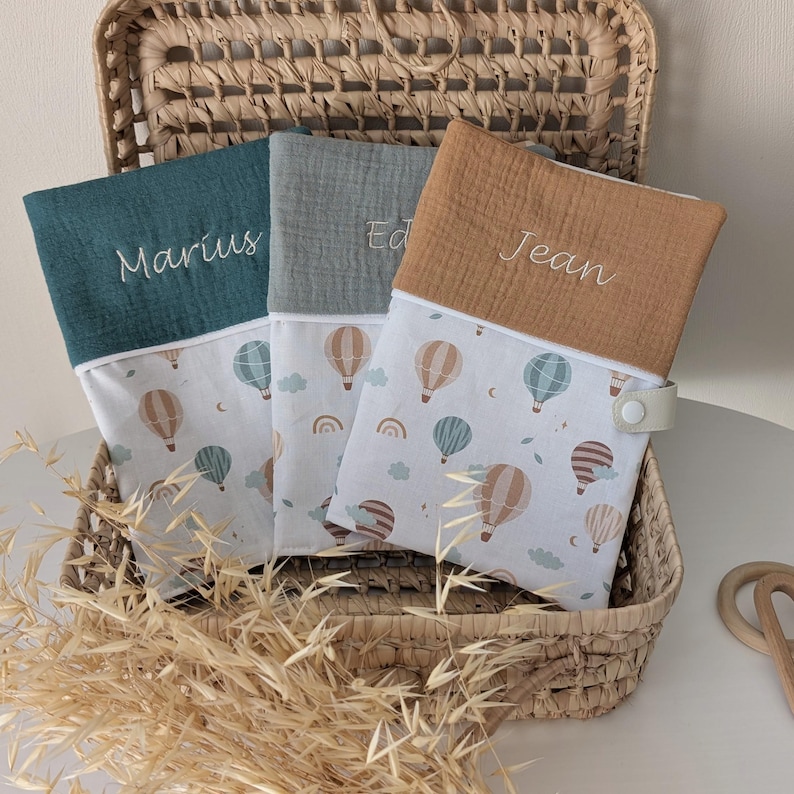 May include: Three personalized baby changing mats in a wicker basket. Each mat has a different color top: teal, gray, and tan. The mats are embroidered with the names "Marius", "Ed", and "Jean". The bottom portion features a hot air balloon pattern.