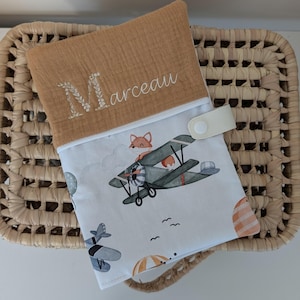 May include: A tan and white baby changing pad with a whimsical airplane and fox print. The pad features the name "Marceau" embroidered in white, and a snap closure. The pad is resting on a woven basket.