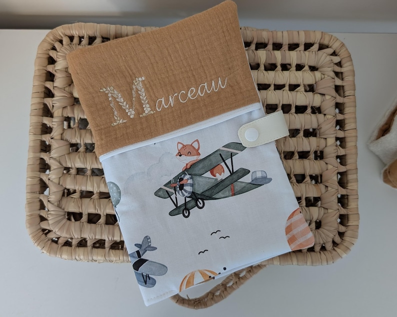 May include: A tan and white baby changing pad with a whimsical airplane and fox print. The pad features the name "Marceau" embroidered in white, and a snap closure. The pad is resting on a woven basket.