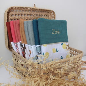 May include: A woven basket filled with a variety of colorful baby blankets. The blankets are in shades of red, pink, orange, and teal, with patterned and solid designs. One teal blanket has the name "Zo&eacute;" embroidered in gold thread. The basket is lined with a lemon-patterned fabric.
