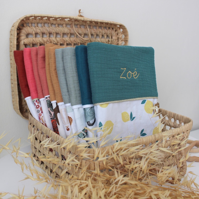 May include: A woven basket filled with a variety of colorful baby blankets. The blankets are in shades of red, pink, orange, and teal, with patterned and solid designs. One teal blanket has the name "Zo&eacute;" embroidered in gold thread. The basket is lined with a lemon-patterned fabric.