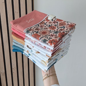 May include: A stack of colorful baby bibs. The top bib is coral with the name "Iris" embroidered in white. The bibs feature floral and patterned designs in various colors, including orange, blue, and yellow. A white snap closure is visible.