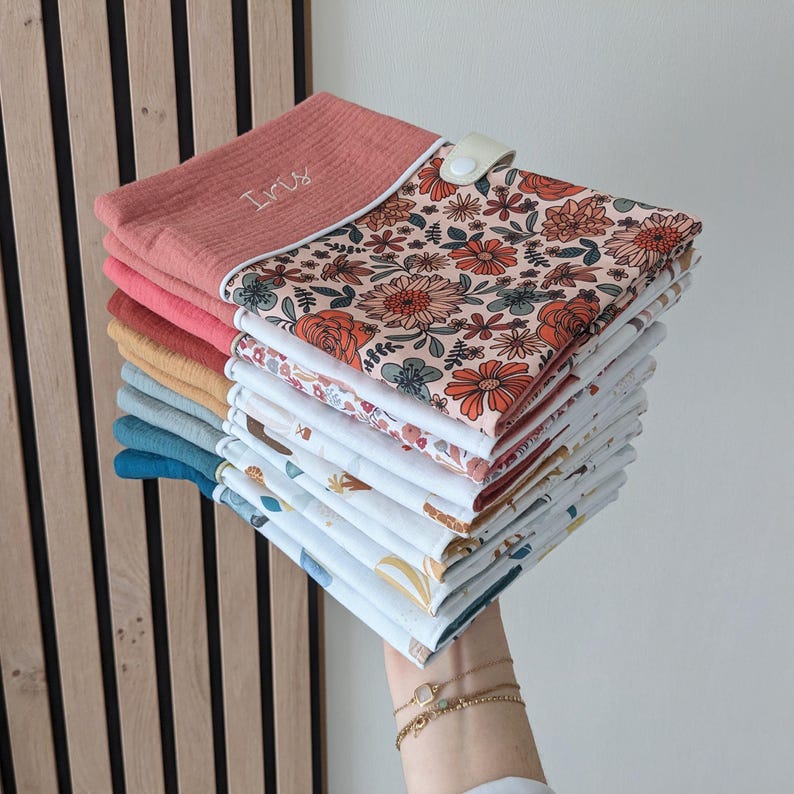 May include: A stack of colorful baby bibs. The top bib is coral with the name "Iris" embroidered in white. The bibs feature floral and patterned designs in various colors, including orange, blue, and yellow. A white snap closure is visible.