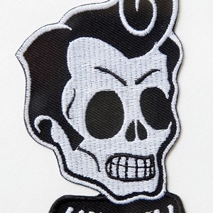 Könnte beinhalten: Gestickter Aufnäher mit einem stilisierten Totenkopf mit Pompadour-Frisur. Der Totenkopf ist weiß mit schwarzen Details und schwarzer Umrandung. Das Wort "KINGLY" ist in Weiß auf einem schwarzen Banner darunter gestickt.