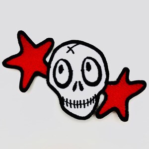 Könnte beinhalten: Ein weißer Cartoon-Schädel mit großen Augen und einem genähten Mund, umgeben von zwei roten fünfzackigen Sternen.