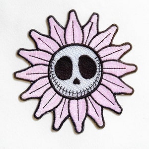 Könnte beinhalten: Rosa Blume mit einem schwarz-weißen Totenkopf im Zentrum. Die Blume hat ein gesticktes Lächeln und ist von rosa Blütenblättern umgeben.