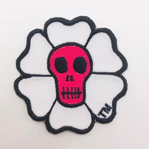 Könnte beinhalten: Ein weißer, blumenförmiger Patch mit einem roten Totenkopf in der Mitte. Der Totenkopf hat schwarze Augen und einen schwarzen Mund. Der Patch hat einen schwarzen Umriss und den Text "TM" in der unteren rechten Ecke.