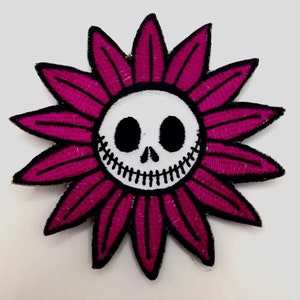 Könnte beinhalten: Eine rosa Blume mit einem weißen Totenkopf im Zentrum. Der Totenkopf hat schwarze Augen und ein genähtes Lächeln.