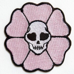 Könnte beinhalten: Rosa gestickter Blumen-Patch mit einem weißen Totenkopf in der Mitte. Der Totenkopf hat schwarze Augen und einen kleinen Mund.