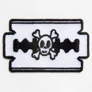 Könnte beinhalten: Ein weißer gestickter Aufnäher in Form einer Rasierklinge mit einem schwarzen Totenkopf- und Knochenmotiv.