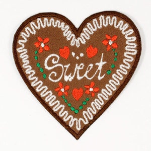 Könnte beinhalten: Ein brauner herzförmiger Lebkuchen mit weißen Nähten an den Rändern. Der Lebkuchen ist mit roten und grünen Stickereien verziert und das Wort "Sweet" in weißer Stickerei.