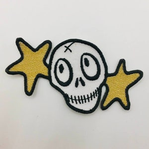 Könnte beinhalten: Weiß gestickter Schädel mit schwarzem Umriss und zwei goldenen Sternen auf jeder Seite. Der Schädel hat ein Kreuz auf der Stirn und zwei große Augen.