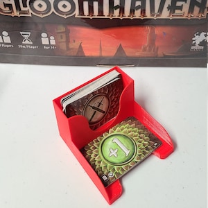 Puede incluir: Porta tarjetas de plástico rojo con dos ranuras para tarjetas. La ranura superior contiene una pila de tarjetas con un símbolo de espadas cruzadas. La ranura inferior contiene una sola tarjeta con un fondo verde y un símbolo "1+" blanco.