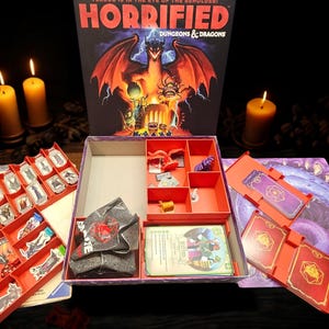 May include: Open board game box with the title "Horrified: Dungeons & Dragons" featuring a dragon illustration. The box contains game pieces, cards, and a game board. Lit candles provide a warm glow.