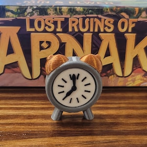 Könnte beinhalten: Ein grauer Wecker mit goldenen Akzenten und einem weißen Zifferblatt. Der Wecker steht auf einer Holzoberfläche. Der Wecker ist eine Spielfigur für das Brettspiel "Lost Ruins of Arnak".