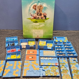 Op de afbeelding: Bordspel "Ark Nova" met een doos met een olifant, beer en andere dieren. Het spel bevat blauwe trays, spelstukken en zeshoekige tegels. De doos is groen en wit.