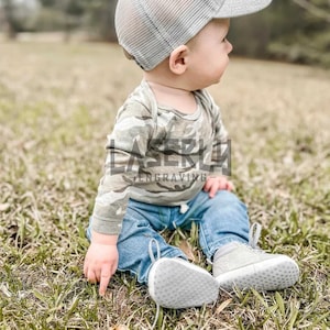 Puede incluir: Un beb&eacute; con una camisa de manga larga de camuflaje, pantalones vaqueros azules y una gorra de camionero de malla gris con un parche marr&oacute;n. El beb&eacute; est&aacute; sentado en un parche de hierba verde.