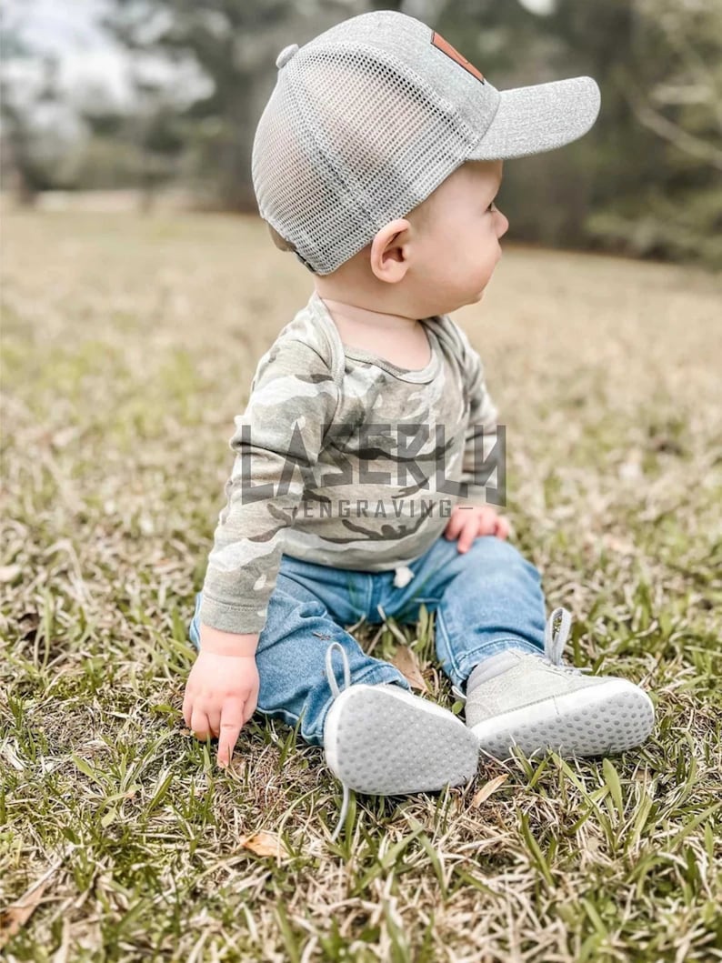 Puede incluir: Un beb&eacute; con una camisa de manga larga de camuflaje, pantalones vaqueros azules y una gorra de camionero de malla gris con un parche marr&oacute;n. El beb&eacute; est&aacute; sentado en un parche de hierba verde.