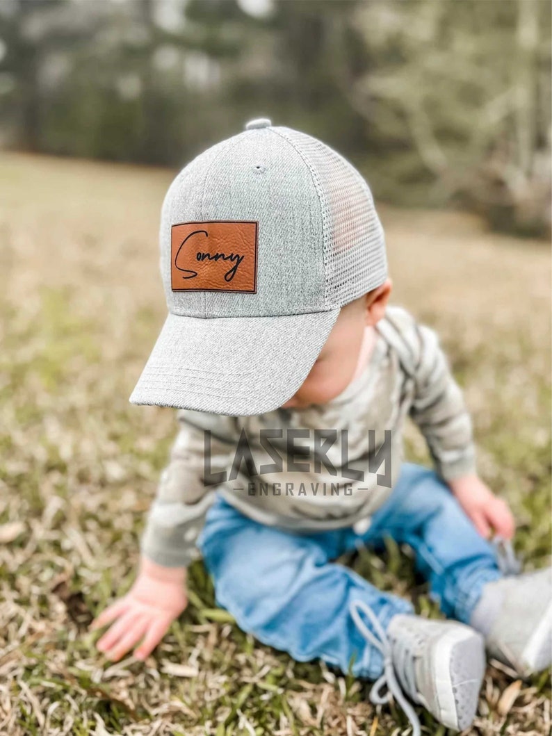 Puede incluir: Una gorra de b&eacute;isbol de malla gris y negra con un parche de cuero marr&oacute;n que dice "Sonny" en cursiva.
