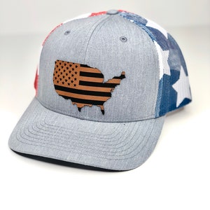 Könnte beinhalten: Eine graue und blaue Trucker-Kappe mit Netzrücken. Die Vorderseite der Kappe zeigt eine braune Lederumrandung der Vereinigten Staaten mit einem schwarz-weißen amerikanischen Flaggenmuster.