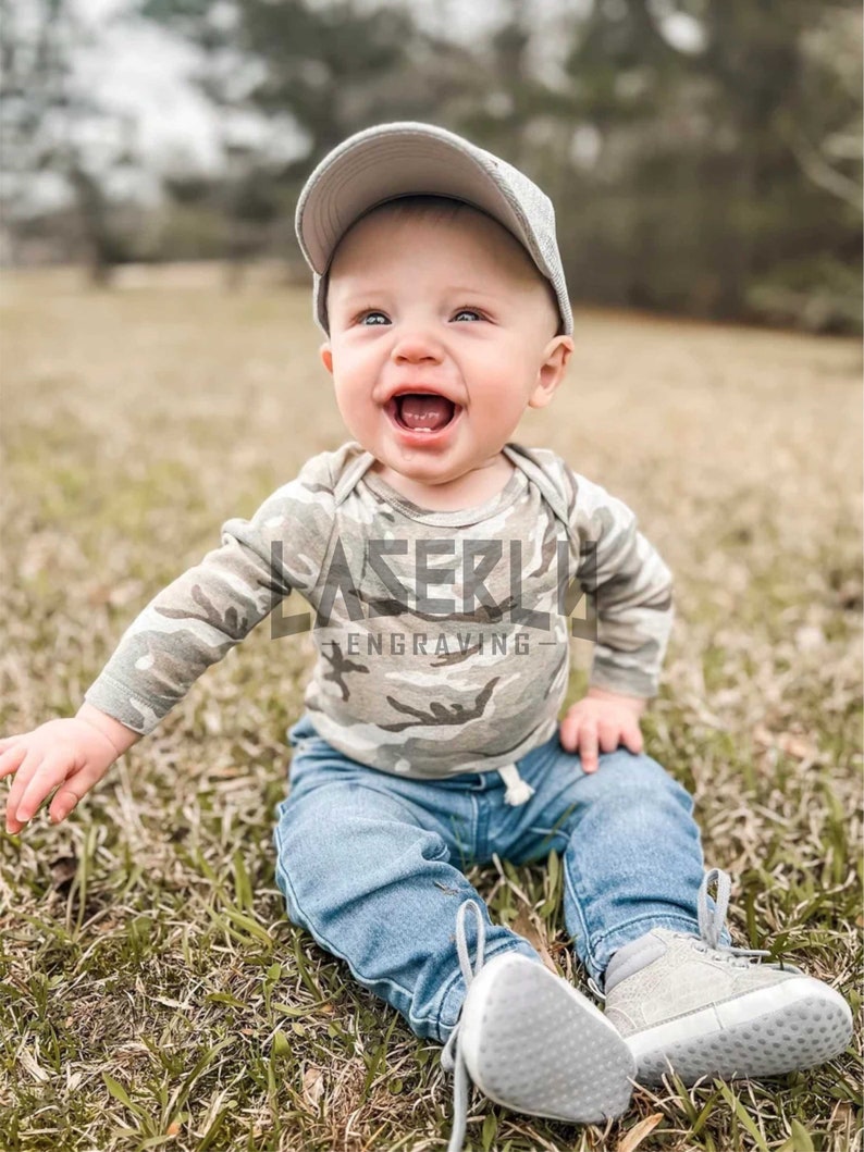 Puede incluir: Un beb&eacute; var&oacute;n con una camisa de manga larga de estampado de camuflaje, pantalones vaqueros azules y una gorra de b&eacute;isbol gris. La camisa tiene el texto "LASER" y "ENGRAVING". El beb&eacute; est&aacute; sentado en un parche de hierba verde.