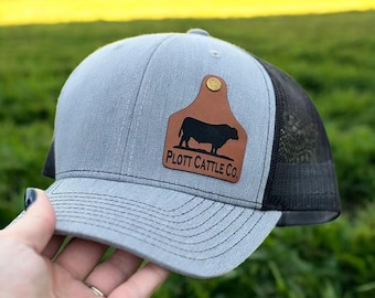 Op de afbeelding: Een grijze en zwarte truckercap met een bruin leren patch met een silhouet van een stier en de tekst "PLOTT CATTLE CO."