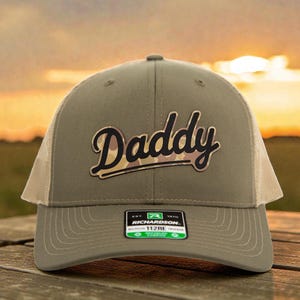 Könnte beinhalten: Khaki-farbene Trucker-Cap mit Mesh-Rückseite und dem Wort "Daddy" in Schreibschrift mit Camouflage-Füllung. Die Kappe hat einen gebogenen Schirm und ein Richardson 112RE-Etikett.