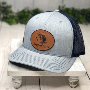 May include: A grey and navy blue trucker hat with a brown leather patch featuring a bass fish and the text "Rippin' Lips".