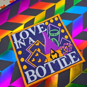 May include: Embroidered patch with the text "LOVE IN A BOTTLE" in white on a purple background. The patch features a bottle, dice, and hearts. The patch is outlined in orange and set against a rainbow geometric pattern.