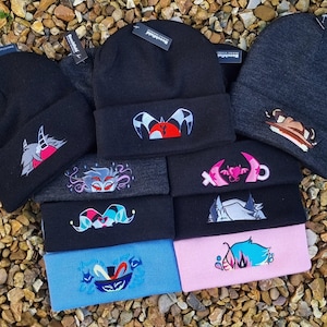 May include: A stack of black, grey and pink knitted beanies with various embroidered designs. The designs include a heart with wings, a jester, a creature with horns, and a face with a crown. The beanies are all made of a soft, comfortable material and are perfect for keeping your head warm in the winter.
