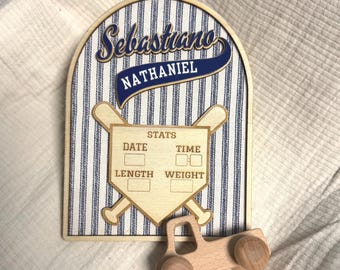 以下が含まれることがあります： 青と白のピンストライプの背景、SebastianoとNATHANIELという名前、日付、時間、長さ、体重の記入欄がある野球のホームベースのグラフィックが描かれたアーチ型の木製プレート。手前には小さな木製のトラックのおもちゃが置かれています。