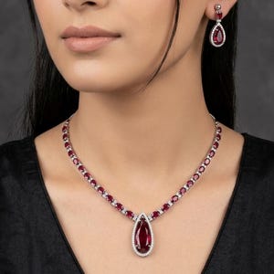 Puede incluir: Un conjunto de collar y pendientes de color rojo rub&iacute; y plata. El collar presenta un gran colgante de rub&iacute; en forma de l&aacute;grima, rodeado de peque&ntilde;as piedras blancas. Los pendientes a juego tienen un dise&ntilde;o similar. La joya se lleva sobre un top negro.