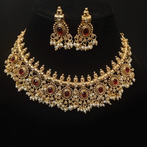 May include: A gold-toned choker necklace and matching drop earrings set featuring oval-shaped red stones, intricate metal filigree detailing, and small white bead accents hanging from the lower edges of the jewelry pieces.