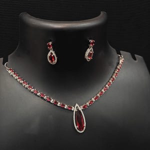 Puede incluir: Un conjunto de collar y pendientes plateados con piedras preciosas rojas en forma de l&aacute;grima. El collar presenta una fila de peque&ntilde;as piedras rojas y un colgante en forma de l&aacute;grima m&aacute;s grande. Los pendientes reflejan el dise&ntilde;o del colgante. La joyer&iacute;a se exhibe en un maniqu&iacute; negro.