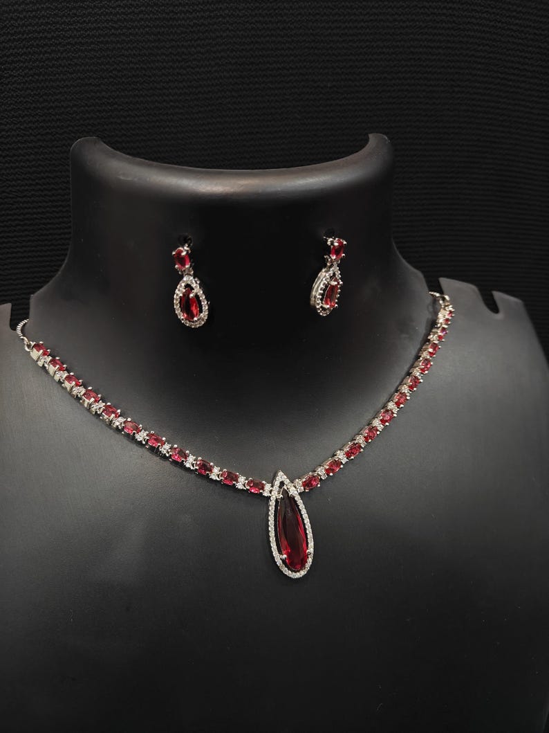 Puede incluir: Un conjunto de collar y pendientes plateados con piedras preciosas rojas en forma de l&aacute;grima. El collar presenta una fila de peque&ntilde;as piedras rojas y un colgante en forma de l&aacute;grima m&aacute;s grande. Los pendientes reflejan el dise&ntilde;o del colgante. La joyer&iacute;a se exhibe en un maniqu&iacute; negro.