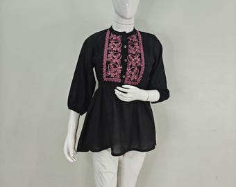 Pode incluir: Top túnica preto com bordado floral rosa na frente. O top tem gola redonda, mangas três quartos e bainha ligeiramente rodada. A túnica é combinada com calças brancas.