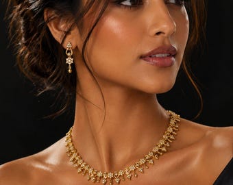 Puede incluir: Un conjunto de collar y pendiente de oro con intrincados detalles metálicos de inspiración floral y pequeñas piedras blancas. El collar se sitúa en la base del cuello, mientras que el pendiente presenta un diseño colgante con forma de lágrima.
