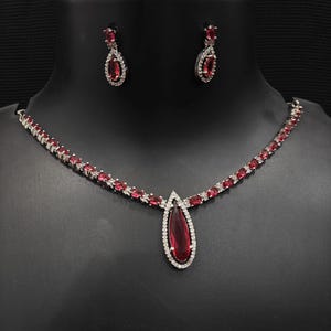 Puede incluir: Un conjunto de joyas plateadas con un collar y pendientes a juego. El collar tiene un colgante en forma de l&aacute;grima con una gran piedra preciosa roja rodeada de piedras blancas m&aacute;s peque&ntilde;as. Los pendientes tambi&eacute;n tienen piedras preciosas rojas y detalles de piedras blancas.