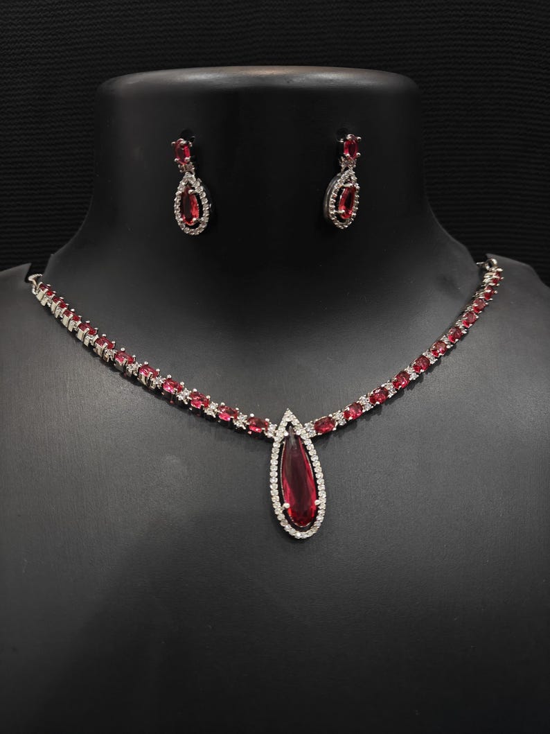 Puede incluir: Un conjunto de joyas plateadas con un collar y pendientes a juego. El collar tiene un colgante en forma de l&aacute;grima con una gran piedra preciosa roja rodeada de piedras blancas m&aacute;s peque&ntilde;as. Los pendientes tambi&eacute;n tienen piedras preciosas rojas y detalles de piedras blancas.