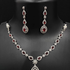 May include: A silver-toned jewelry set featuring a necklace and matching earrings. The necklace and earrings have a teardrop design with a large, red gemstone at the center, surrounded by smaller, clear stones. The set is displayed on a black mannequin.