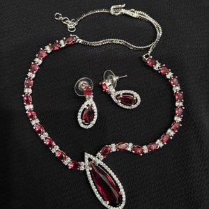 Puede incluir: Un conjunto de collar y pendientes plateados con piedras preciosas rojas rub&iacute; en forma de l&aacute;grima y ovalada. El collar y los pendientes est&aacute;n adornados con peque&ntilde;os cristales transparentes. El conjunto de joyas se muestra sobre una superficie negra.