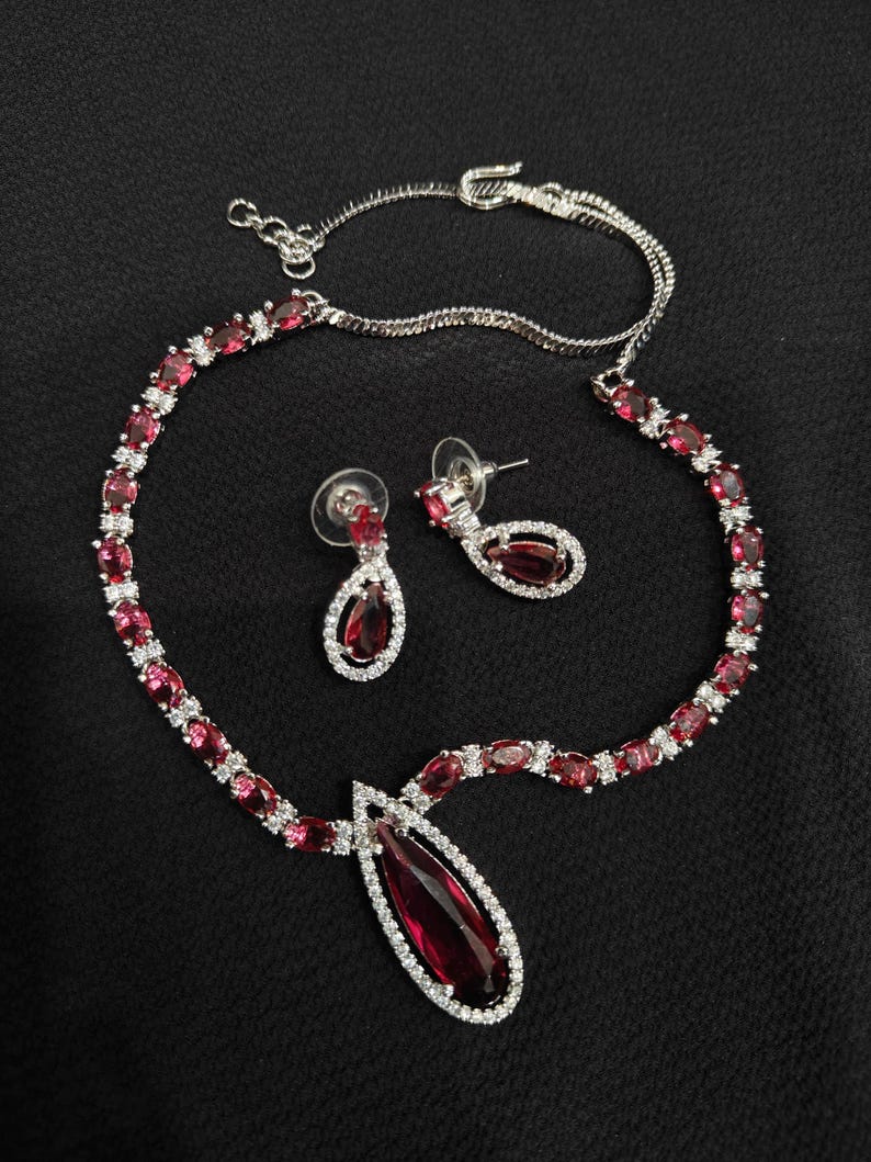 Puede incluir: Un conjunto de collar y pendientes plateados con piedras preciosas rojas rub&iacute; en forma de l&aacute;grima y ovalada. El collar y los pendientes est&aacute;n adornados con peque&ntilde;os cristales transparentes. El conjunto de joyas se muestra sobre una superficie negra.