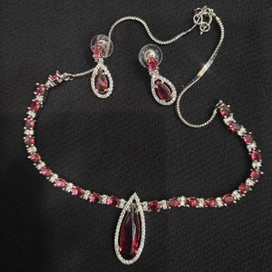 Puede incluir: Un conjunto de joyas plateadas que incluye un collar y pendientes a juego. El collar presenta un colgante en forma de l&aacute;grima y est&aacute; adornado con piedras preciosas rojas y cristales transparentes. Los pendientes tambi&eacute;n tienen un dise&ntilde;o de l&aacute;grima.