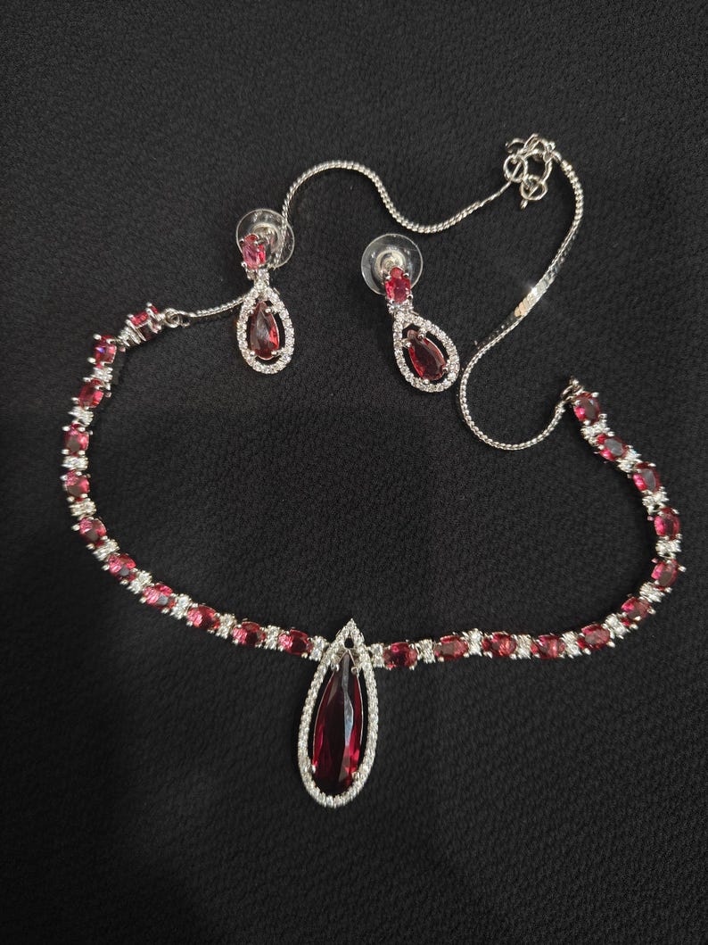 Puede incluir: Un conjunto de joyas plateadas que incluye un collar y pendientes a juego. El collar presenta un colgante en forma de l&aacute;grima y est&aacute; adornado con piedras preciosas rojas y cristales transparentes. Los pendientes tambi&eacute;n tienen un dise&ntilde;o de l&aacute;grima.