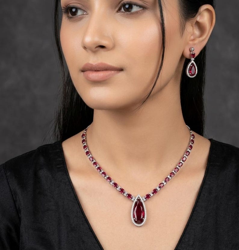 Puede incluir: Un conjunto de collar y pendientes de color rojo rub&iacute; y plata. El collar presenta un gran colgante de rub&iacute; en forma de l&aacute;grima, rodeado de peque&ntilde;as piedras blancas. Los pendientes a juego tienen un dise&ntilde;o similar. La joya se lleva sobre un top negro.