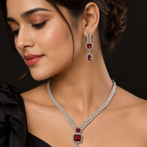 May include: A silver-toned necklace and earring set featuring clear pavé-set stones arranged in a leaf-like pattern. The necklace includes a central pendant with a square red stone and a teardrop red stone. The earrings match the pendant design.