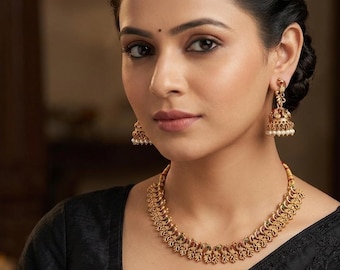 May include: A gold-toned necklace and matching earrings set. The necklace features a detailed, curved design with small red and green accents. The earrings have a bell shape with dangling pearls. The jewelry is worn with a black top.