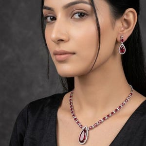 Puede incluir: Un conjunto de collar y pendientes de color rojo rub&iacute;. El collar presenta un colgante en forma de l&aacute;grima y piedras ovaladas. Los pendientes tienen un dise&ntilde;o similar en forma de l&aacute;grima. Ambas piezas est&aacute;n engastadas en un metal plateado y son llevadas por una persona.