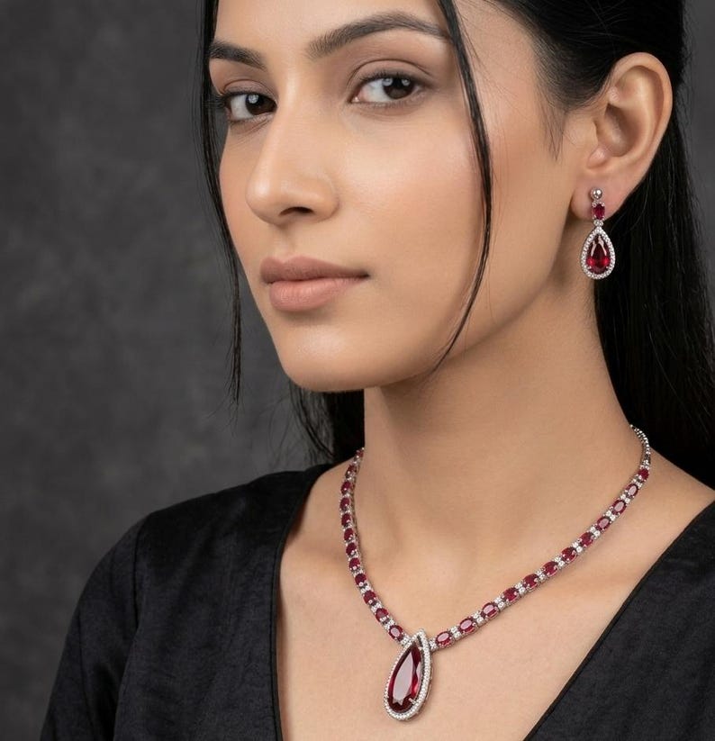 Puede incluir: Un conjunto de collar y pendientes de color rojo rub&iacute;. El collar presenta un colgante en forma de l&aacute;grima y piedras ovaladas. Los pendientes tienen un dise&ntilde;o similar en forma de l&aacute;grima. Ambas piezas est&aacute;n engastadas en un metal plateado y son llevadas por una persona.